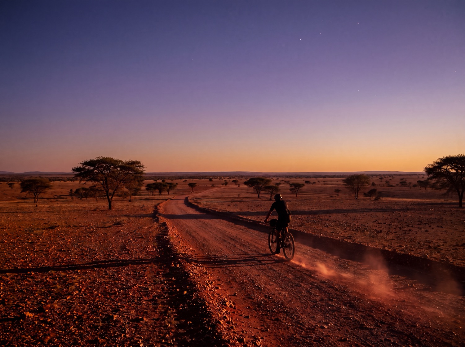 A lone cyclist on a mountain bike rides a red-gravel road at dusk, the African sky turning purple-blue.