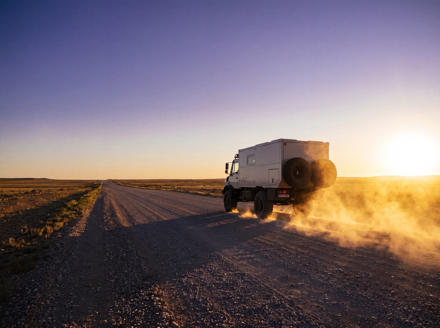 The Unimog driving away down a gravel road into a low golden sun, dust trailing gold behind it.
