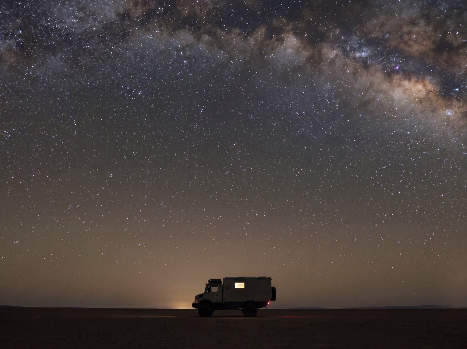 The Unimog parked alone on a salt pan beneath the arch of the Milky Way, one window lit.